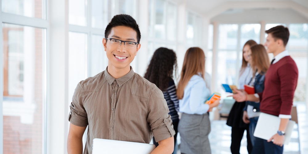Portrait of a cheerful male student standing in university hall with classmates on background